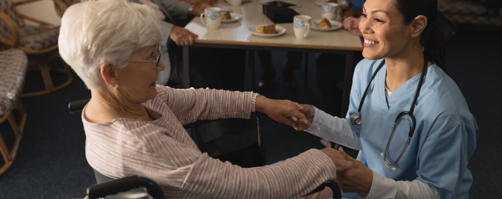 Side view of happy female doctor holding hands and talking with disable senior woman at nursing home